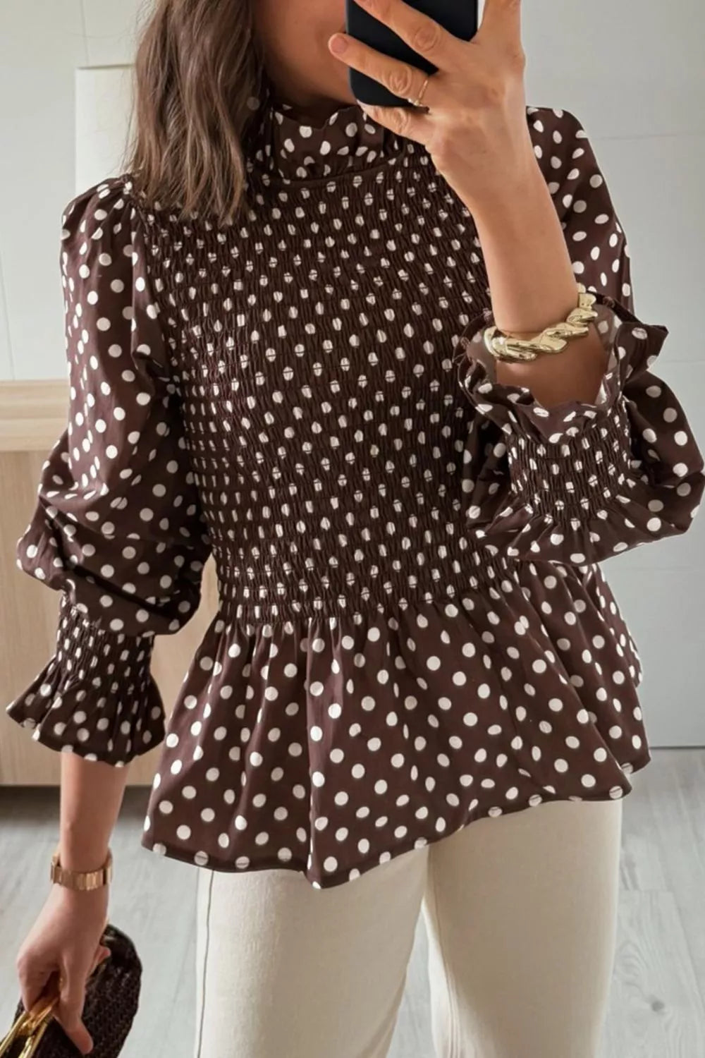 Mujer con blusa marrón de lunares blancos, cintura fruncida, cuello alto y mangas largas abullonadas con volantes. NINA AURA COLLECTION.